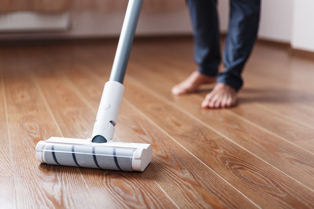 Pernas humanas e uma escova turbo branca de um aspirador sem fio limpam o parquete da casa. tecnologias modernas para limpeza.