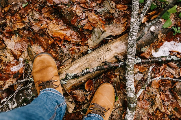 Pernas masculinas com botas marrons em folhas caídas ao lado de um obstáculo de madeira, close-up