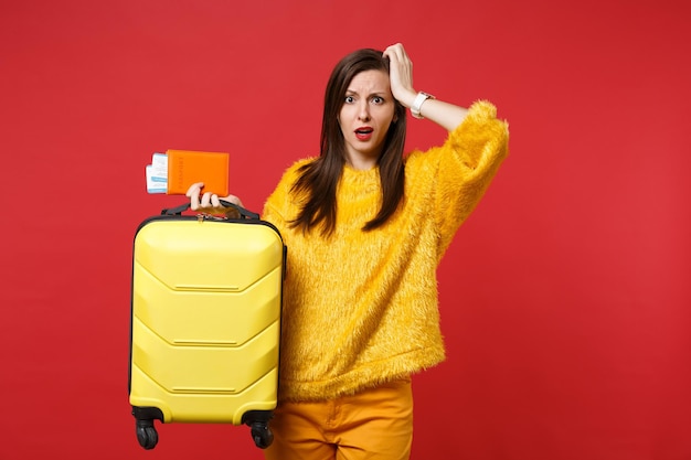 Foto perplejo joven preocupado poniendo la mano en la cabeza, sosteniendo la maleta, pasaporte boleto de embarque aislado sobre fondo rojo brillante. personas sinceras emociones, concepto de estilo de vida. simulacros de espacio de copia.