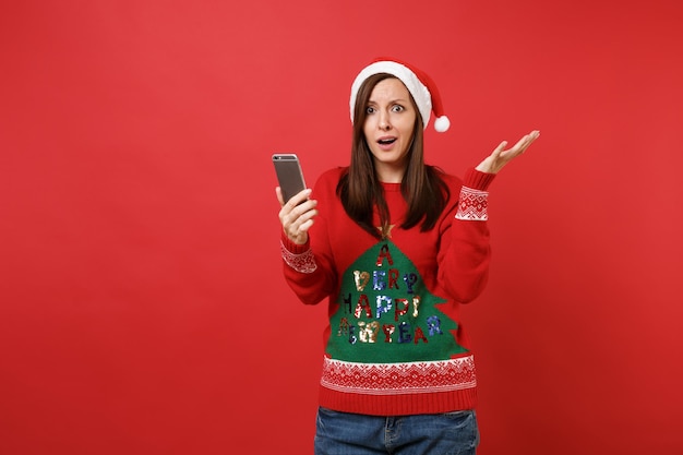 Perplejo joven Santa en suéter de punto, sombrero de Navidad extendiendo las manos sosteniendo el teléfono móvil aislado sobre fondo rojo. Feliz año nuevo 2019 celebración concepto de fiesta navideña. Simulacros de espacio de copia.