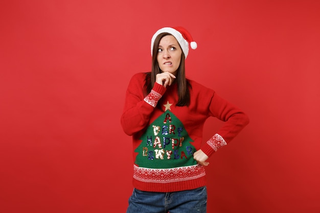 Perplexa pensativa jovem santa no chapéu de natal, olhando para cima, levantou a mão apoiada no queixo mordendo os lábios isolados no fundo vermelho. conceito de festa de feriado de celebração de ano novo feliz 2019. simule o espaço da cópia.