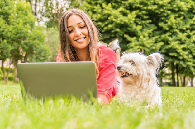 El perro está mirando a su dueño usando la PC. Joven atractiva está estudiando tumbado en el parque con su perro. Concepto sobre perro, personas, naturaleza y tecnología.