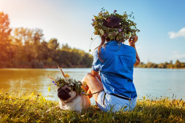 Perro Pug y su maestro relajarse junto al río con coronas de flores. Feliz cachorro y mujer disfrutando de la naturaleza de verano al aire libre
