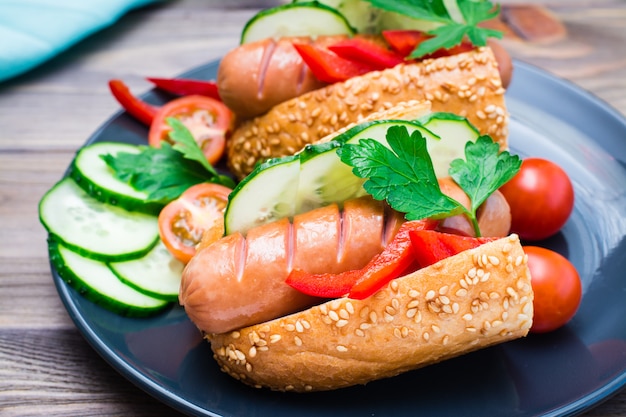 Perros calientes listos para comer de salchichas fritas, bollos de sésamo y verduras frescas en un plato sobre una mesa de madera. De cerca