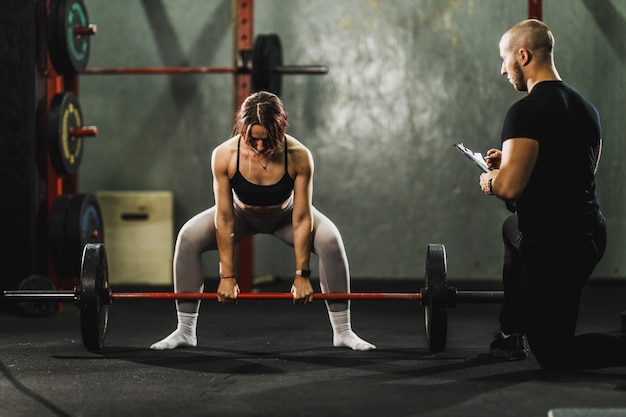 Persönlicher Trainer, der eine muskulöse junge Frau beim Krafttraining im Fitnessstudio beobachtet und unterstützt. Sie macht Kreuzheben mit Langhantel.