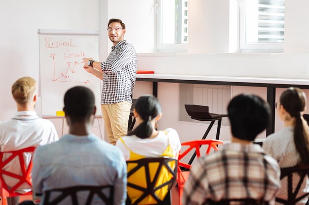 Persönliches Wachstum. Positiver begeisterter Sprecher, der lächelt und auf das Flipchart zeigt, während er vor der Gruppe steht und über persönliches Wachstum spricht