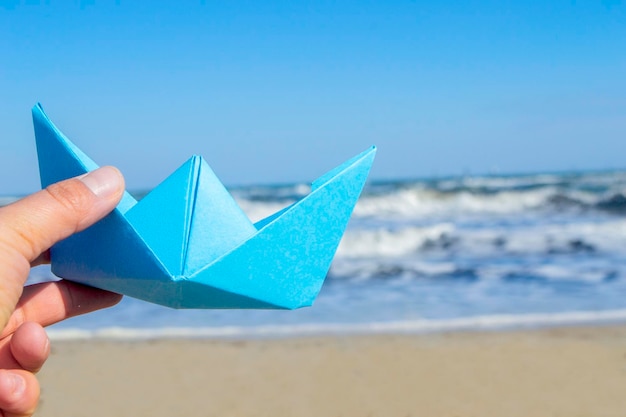 Person, die blaues Papierboot in der Hand auf Hintergrund des blauen Himmels der Meereswellen hält