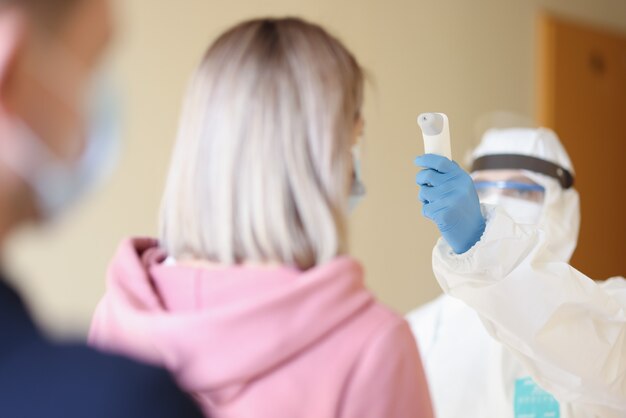 Person, die einen medizinischen Schutzanzug und eine Maske trägt, misst die Temperatur mit einem Thermometer für Besucher