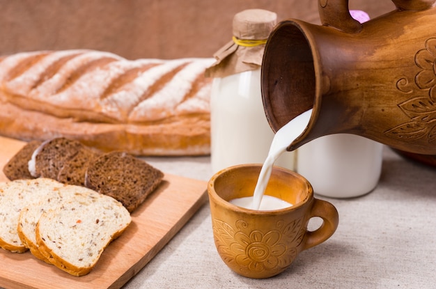 Person, die frische cremige Bauernmilch in einen rustikalen Keramikbecher mit geschnittenem frischem Brot auf einem Brett für ein gesundes Frühstück gießt