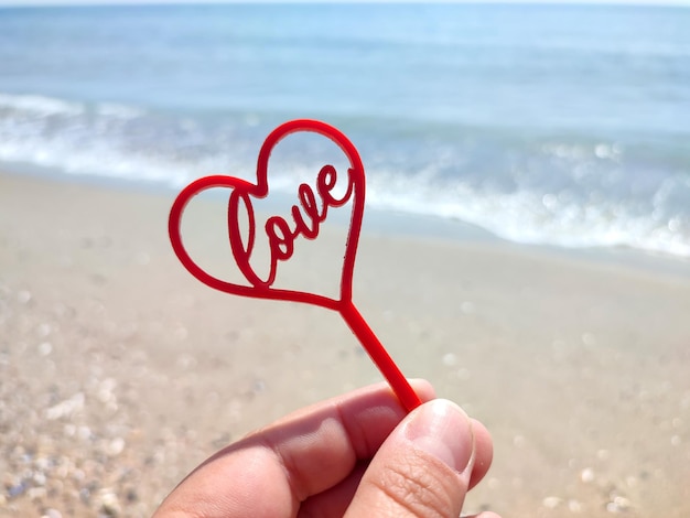 Person hält in den Fingern einen Stock in Form eines roten Herzens und das Wort „Liebe“ am Sandstrand im Hintergrund