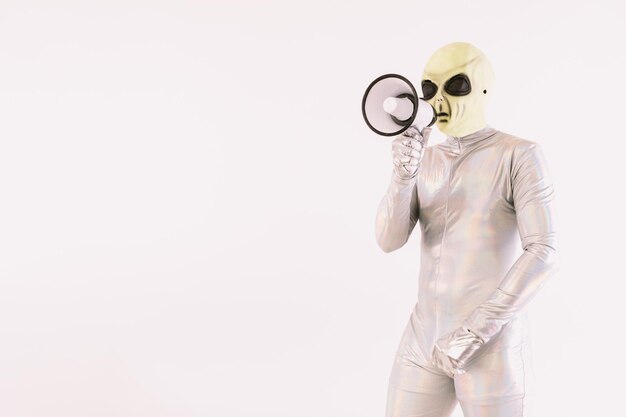 Person in silbernem Anzug und grüner Alien-Maske, die mit einem weißen Megaphon auf weißem Hintergrund spricht