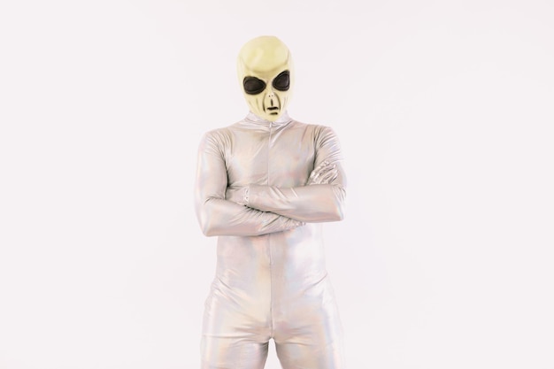 Person in silbernem Anzug und grüner Alien-Maske mit verschränkten Armen, auf weißem Hintergrund