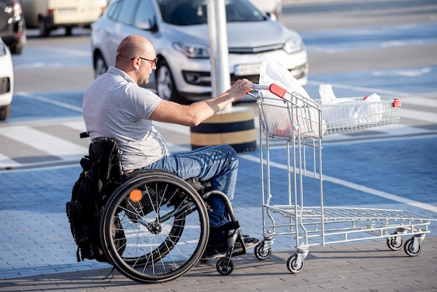 Person mit einer körperlichen Behinderung, die auf einem Supermarktparkplatz einen Einkaufswagen vor sich her schiebt