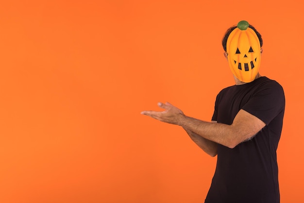 Foto person mit kürbismaske, die halloween feiert und mit den fingern auf orangefarbenem hintergrund zur seite zeigt konzept der feier allerseelen und allerheiligen