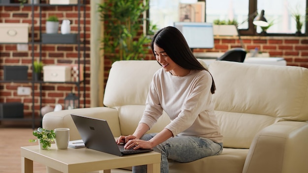 Persona adulta joven que hace trabajo remoto en una computadora portátil moderna mientras está en casa. Mujer sonriente de corazón en casa trabajando remotamente en una laptop mientras se sienta en un sofá en la sala de estar.