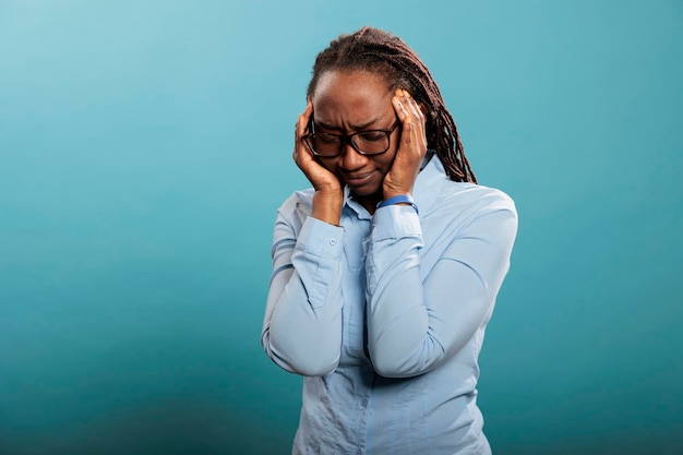 Persona adulta joven sobrecargada de trabajo y estresada que presiona las sienes con los dedos debido al dolor de cabeza. Mujer afroamericana que sufre de migraña de fondo azul.