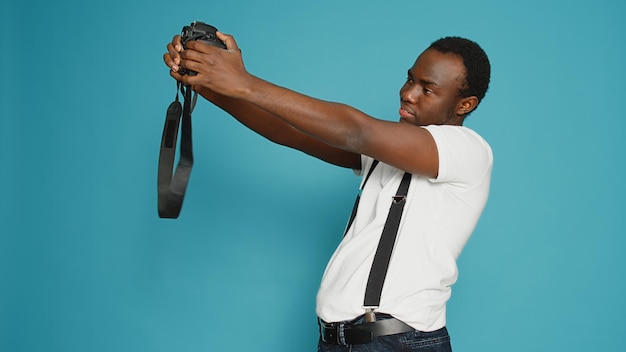 Persona afroamericana sosteniendo una cámara dslr para tomar fotos, de pie en el estudio sobre fondo azul. Fotógrafo que usa equipo profesional para capturar fotos, zoom de enfoque.