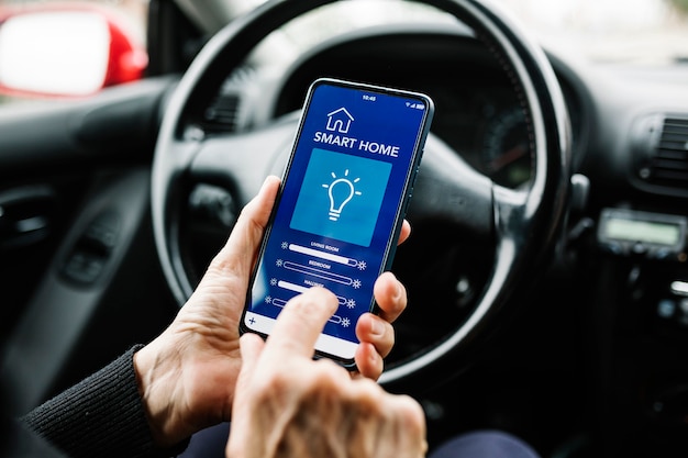 Persona anónima sentada en el asiento del conductor en el automóvil y usando un teléfono móvil con la aplicación de casa inteligente en la pantalla