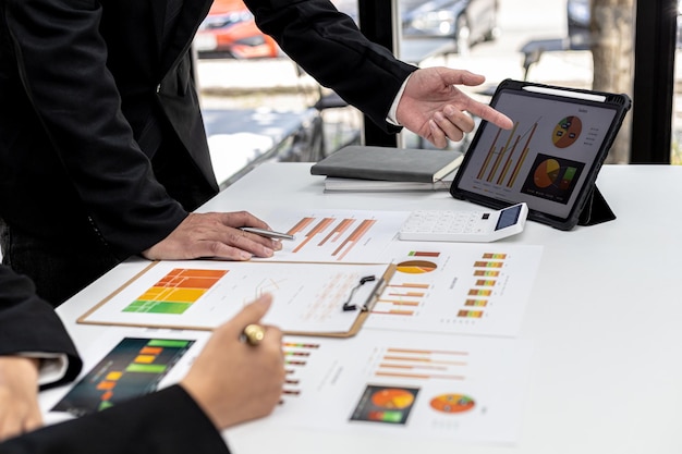 Una persona apunta a la pantalla de una tableta en una pantalla que muestra archivos de datos financieros, dos empresarios intercambian ideas para resolver pérdidas y planificar estrategias para mantener la empresa rentable y en crecimiento.