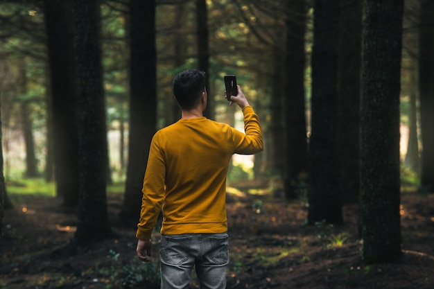 Persona con chaqueta amarilla y jeans grises tomando fotos con el móvil en el bosque