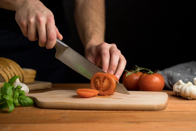 Persona cortando tomate en una tabla de madera preparando un tentempié italiano de bruschetta de fondo oscuro