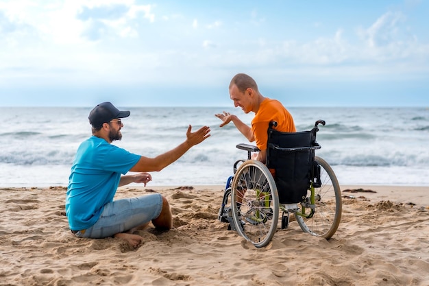 Una persona discapacitada en una silla de ruedas en la playa con un amigo sentado mirando el mar de la mano