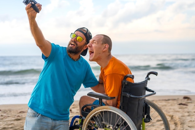 Una persona discapacitada en una silla de ruedas en la playa divirtiéndose con un amigo y una cámara de video tomando una selfie