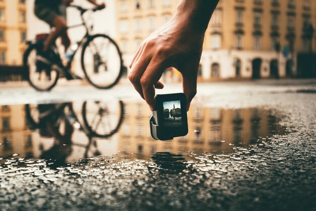 Foto persona filmando a un ciclista desde el nivel de la superficie con el reflejo en la ciudad durante la temporada de lluvias