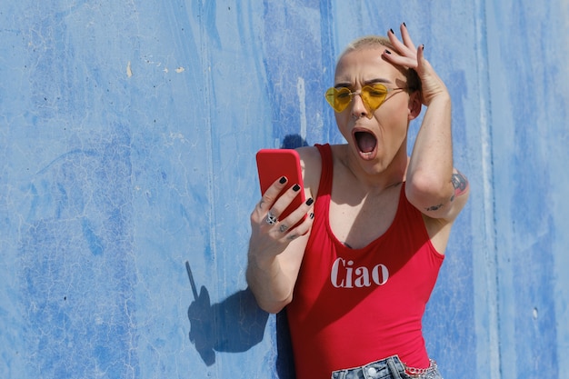 Persona joven no binaria mirando con una expresión de asombro y sorpresa en la pantalla del teléfono móvil mientras lo usa al aire libre.