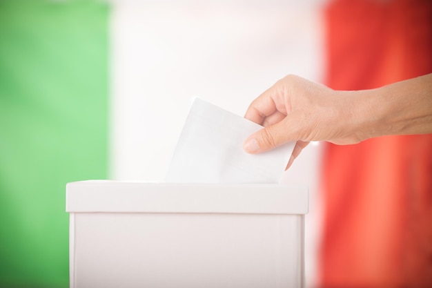 Foto persona de mano poniendo papeleta en una urna durante las elecciones en italia frente a la bandera