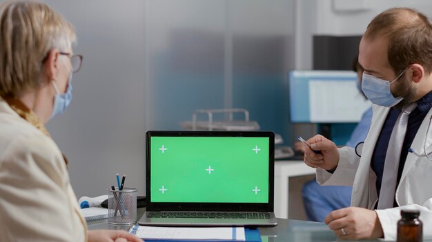 Persona mayor que mira la pantalla verde en la computadora portátil en la visita de control con el médico durante la pandemia. Paciente mayor y médico que analizan el espacio de copia de la maqueta con una plantilla cromakey aislada en el examen.