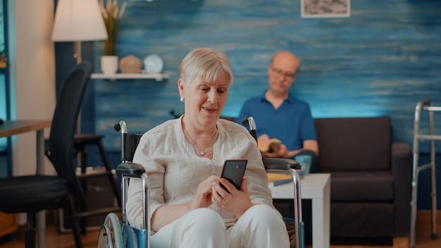 Persona mayor sentada en silla de ruedas y usando un teléfono inteligente para navegar por Internet y descubrir tecnología. Anciana que sufre de discapacidad crónica y mira la pantalla del teléfono móvil.