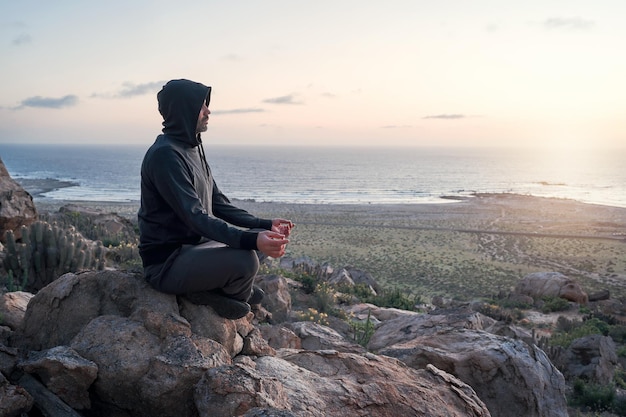 Persona meditando sobre una roca en la colina al atardecer con el océano de fondo en el horizonte