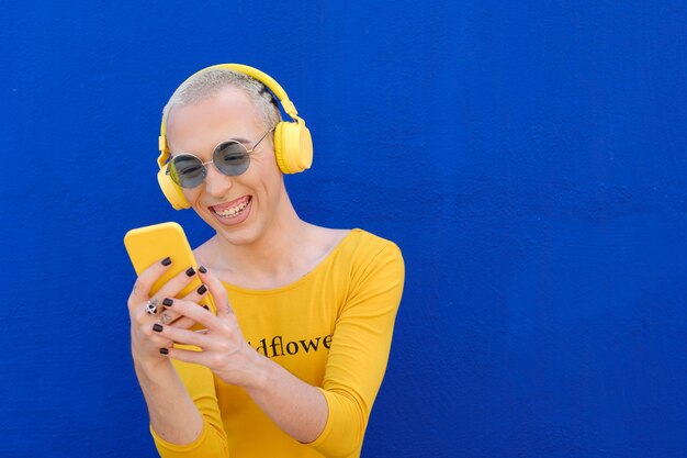 Persona no binaria despreocupada sonriendo mientras usa un teléfono móvil con auriculares inalámbricos al aire libre contra una pared azul.