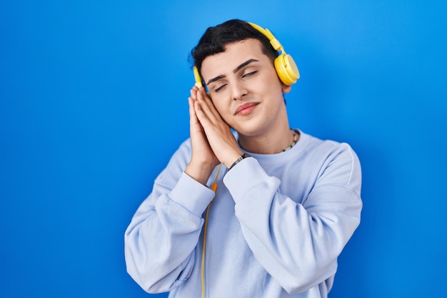 Persona no binaria escuchando música usando auriculares durmiendo cansado soñando y posando con las manos juntas mientras sonríe con los ojos cerrados