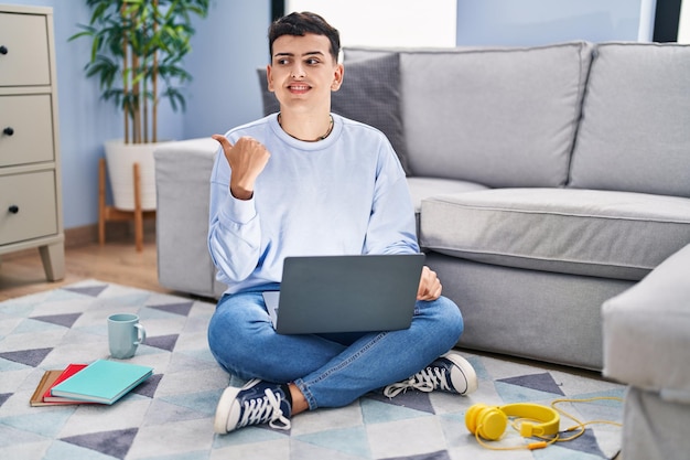 Persona no binaria que estudia usando una computadora portátil sentada en el suelo sonriendo con cara feliz mirando y apuntando hacia un lado con el pulgar hacia arriba