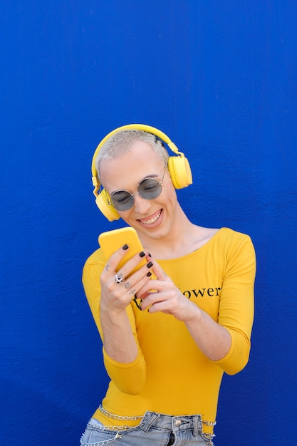 Persona no binaria sonriente usando un teléfono móvil con auriculares inalámbricos al aire libre contra una pared azul. Concepto de identidad de género.