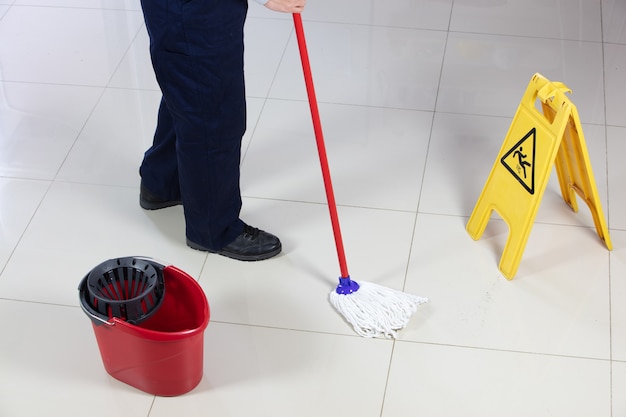 Persona que limpia el piso con un trapeador rojo cerca de un cartel amarillo de piso mojado de precaución