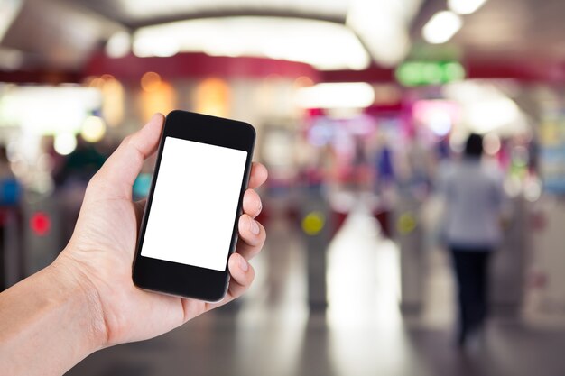 Persona que usa el sostenedor blanco de la pantalla del smartphone en la mano con el fondo enmascarado de la gente que recorre después de máquinas del boleto en estación de tren eléctrica.