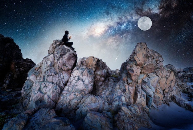 Persona en la roca al aire libre meditando o rezando por la noche bajo la Vía Láctea y la Luna llena