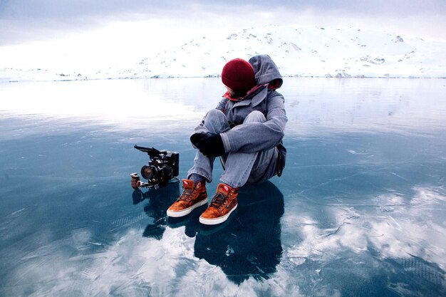 Foto persona sentada en un lago congelado por una montaña cubierta de nieve