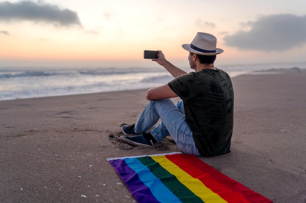 Persona sentada en la orilla de la playa con bandera del arco iris tomando fotos al atardecer LGTBQ