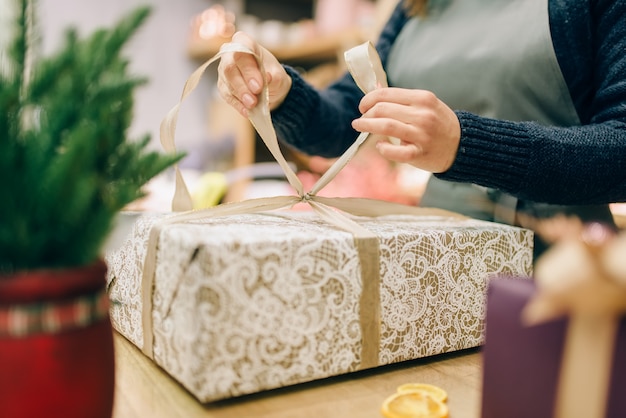 Persona de sexo femenino ata un lazo de oro en caja de regalo, proceso de decoración y envoltura artesanal. Mujer envuelve presente sobre la mesa, procedimiento de decoración