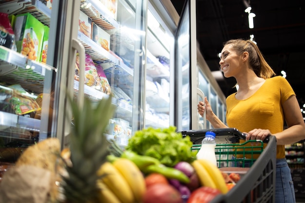 Persona de sexo femenino con carrito de compras y tomando alimentos congelados de la nevera en la tienda de comestibles.