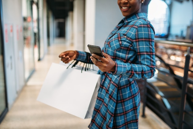 Persona de sexo femenino negro con teléfono y bolsas de compras en el centro comercial. Adicto a las compras en tienda de ropa, estilo de vida de consumismo, moda