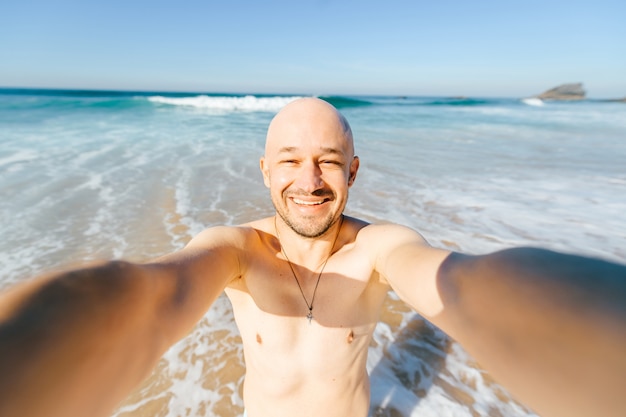 Persona de sexo masculino alegre alegre alegre feliz que hace el selfie en el mar bajo rayos del sol. Hombre alegre de vacaciones después de nadar en el océano de pie en la playa con olas y riéndose de la cámara. Distorsión de la lente.