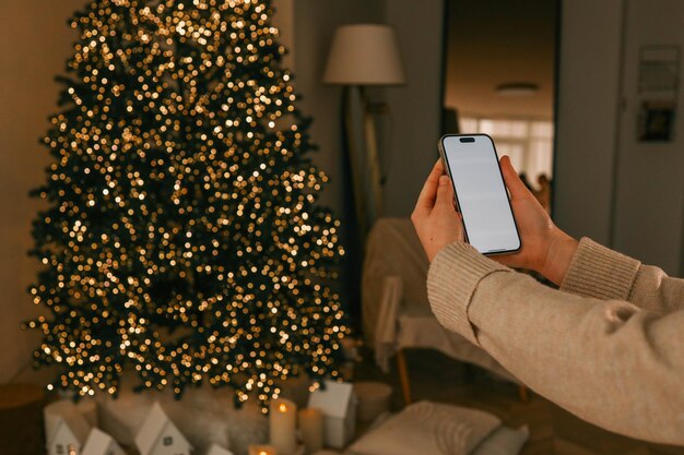 Persona sosteniendo un teléfono inteligente con una pantalla en blanco en un acogedor entorno navideño Un árbol de Navidad decorado brilla cálidamente en el fondo creando una atmósfera festiva y serena