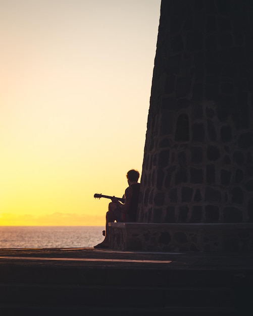 Persona tocando la guitarra sentada en la base de un faro mientras observa el atardecer