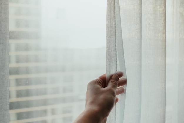 Persona tocando y sosteniendo una cortina blanca en la habitación al lado de la ventana