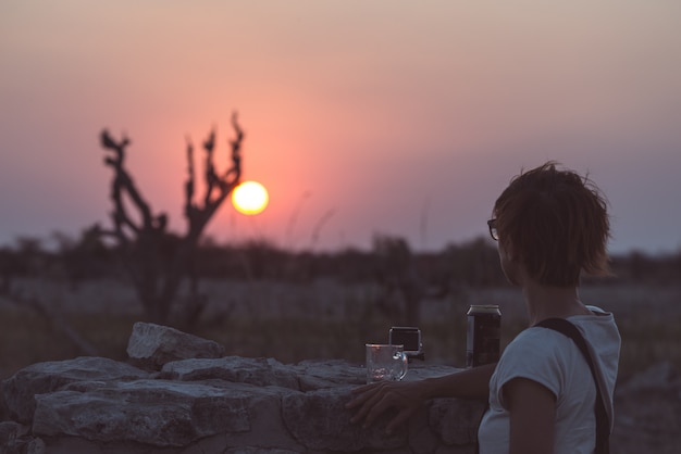 Una persona tomando una copa y mirando la colorida puesta de sol en el desierto de Namib, destino de viaje en Namibia, África. Concepto de aventura y gente viajera.